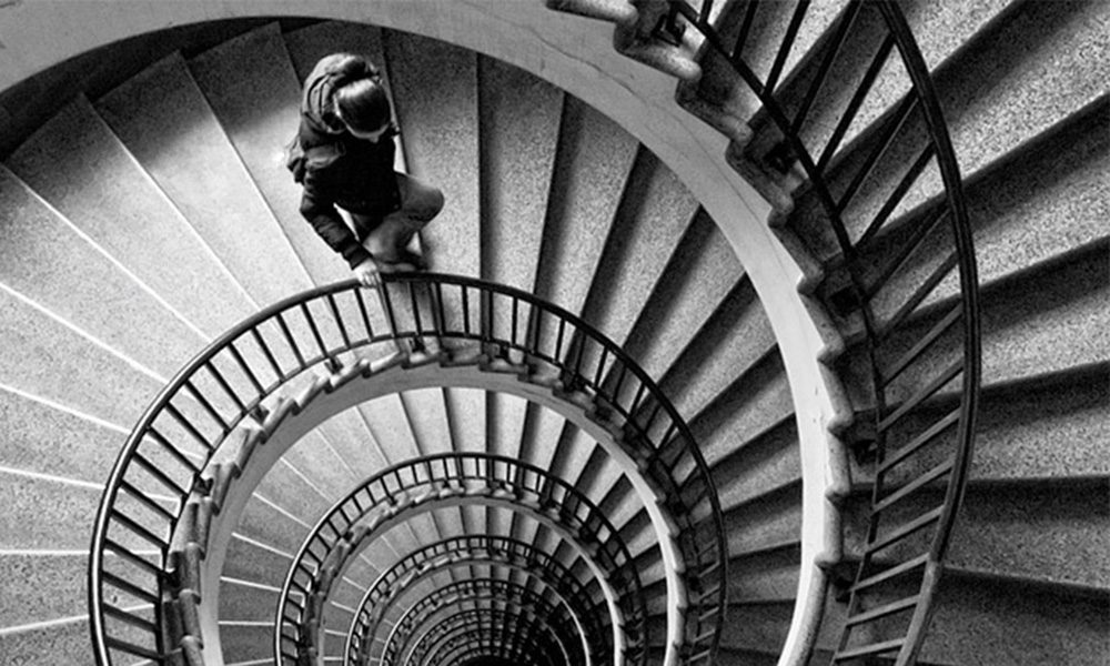Black and white drawing of nondescript person walking down a flight of seemingly endless spiral stairs